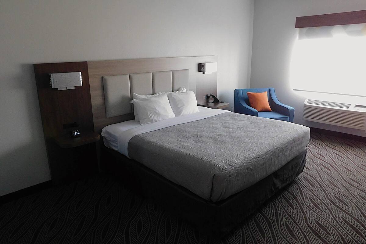 Habitación de hotel con una cama grande, una mesita de noche con un reloj, una silla y una ventana con aire acondicionado.