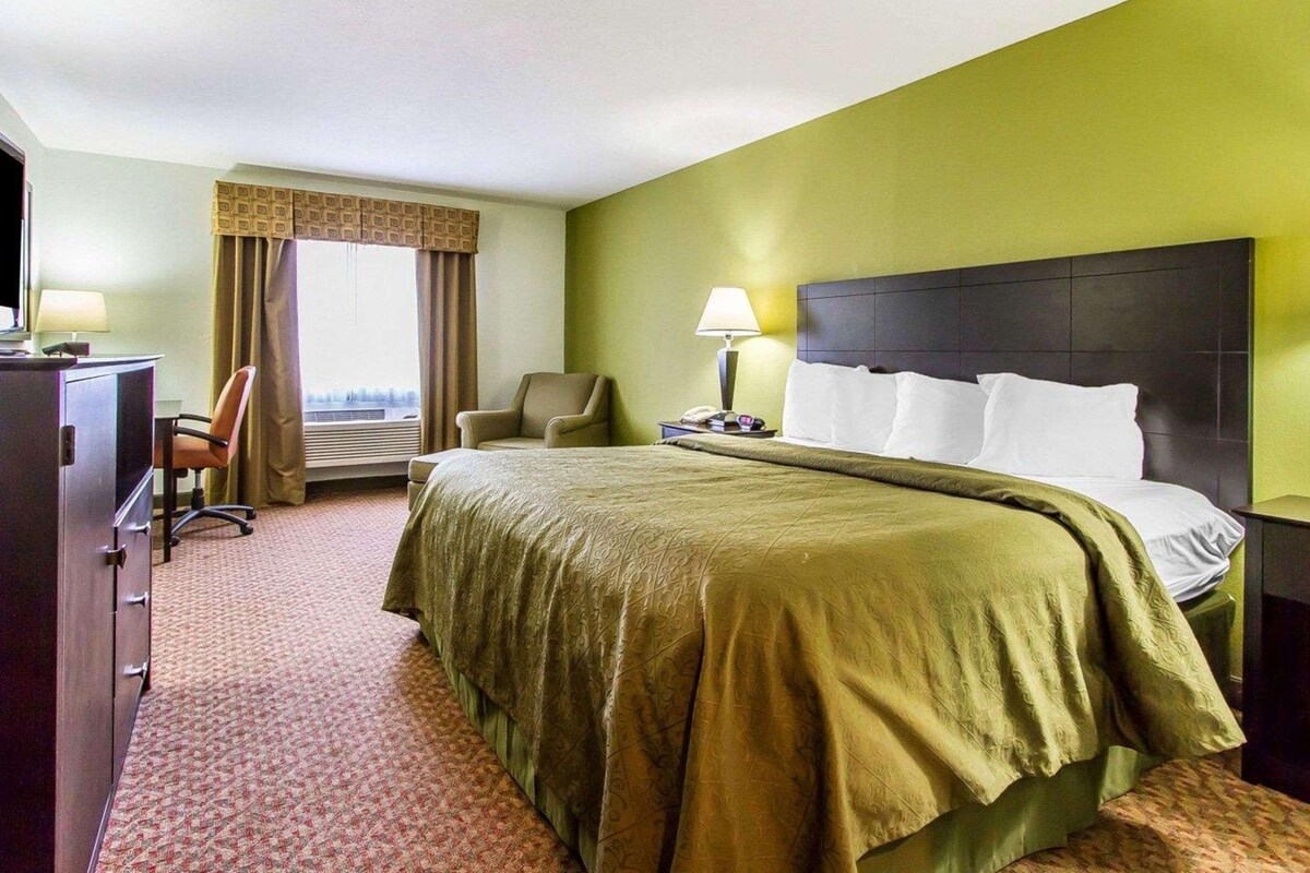 A hotel room with a large bed, a desk with a lamp, a chair, and a view of the city through the window.