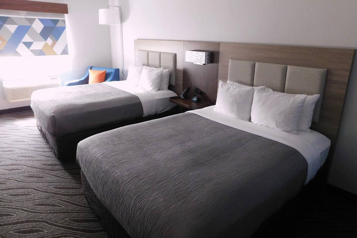 Habitación de hotel con dos camas, una mesita de noche, una lámpara y una ventana con diseño geométrico.