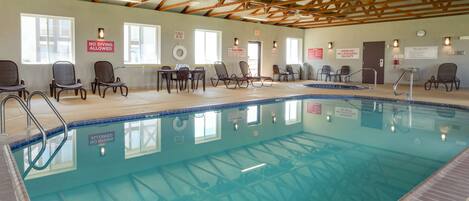 An indoor swimming pool with wooden beams on the ceiling, lounge chairs, and safety signs on the walls.