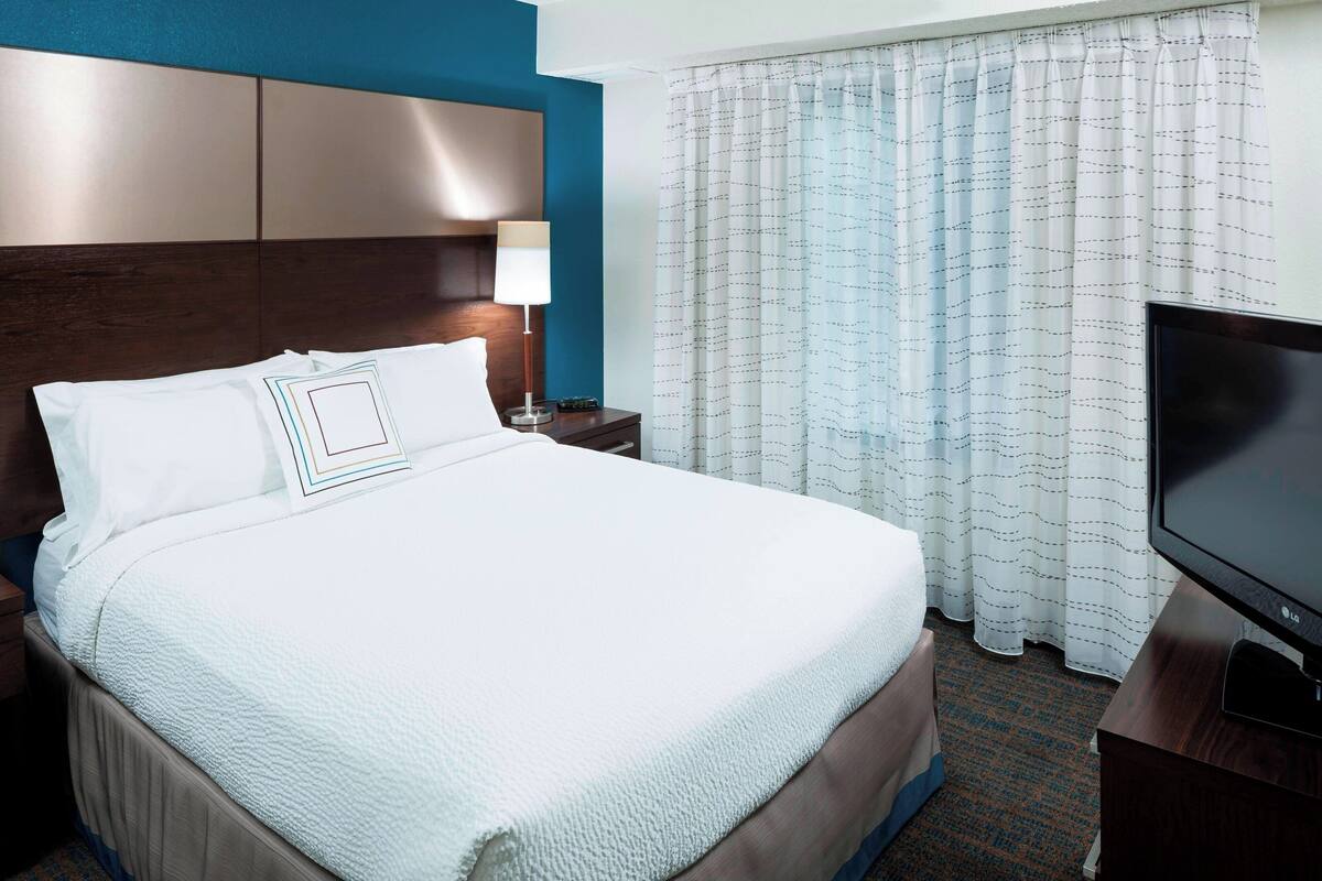 A hotel room with a large bed, a flat-screen TV, a nightstand, and a window with curtains.