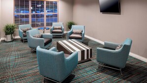 A hotel lobby with a seating area featuring teal armchairs and a striped ottoman, a flat-screen TV mounted on the wall, and a large window with blinds.