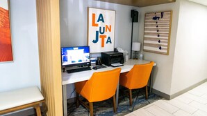 Una habitación de hotel con un escritorio, dos sillas naranjas, una computadora, una impresora y una obra de arte enmarcada en la pared.