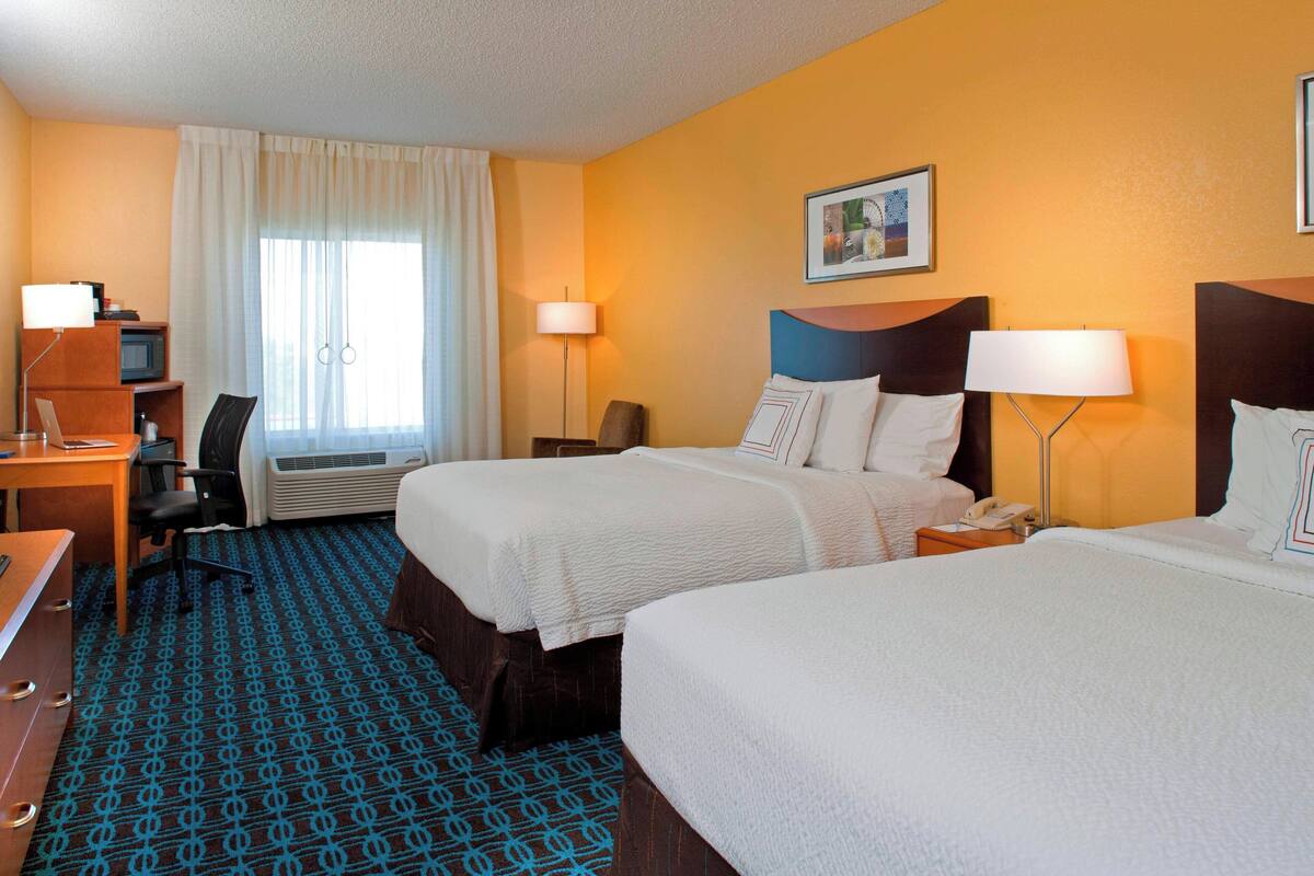 A hotel room with two beds, a desk, a chair, a lamp, and a picture on the wall.