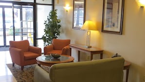 A lobby with two orange armchairs, a round coffee table with a bowl, a green sofa, a potted plant, a lamp, and a framed picture on the wall.