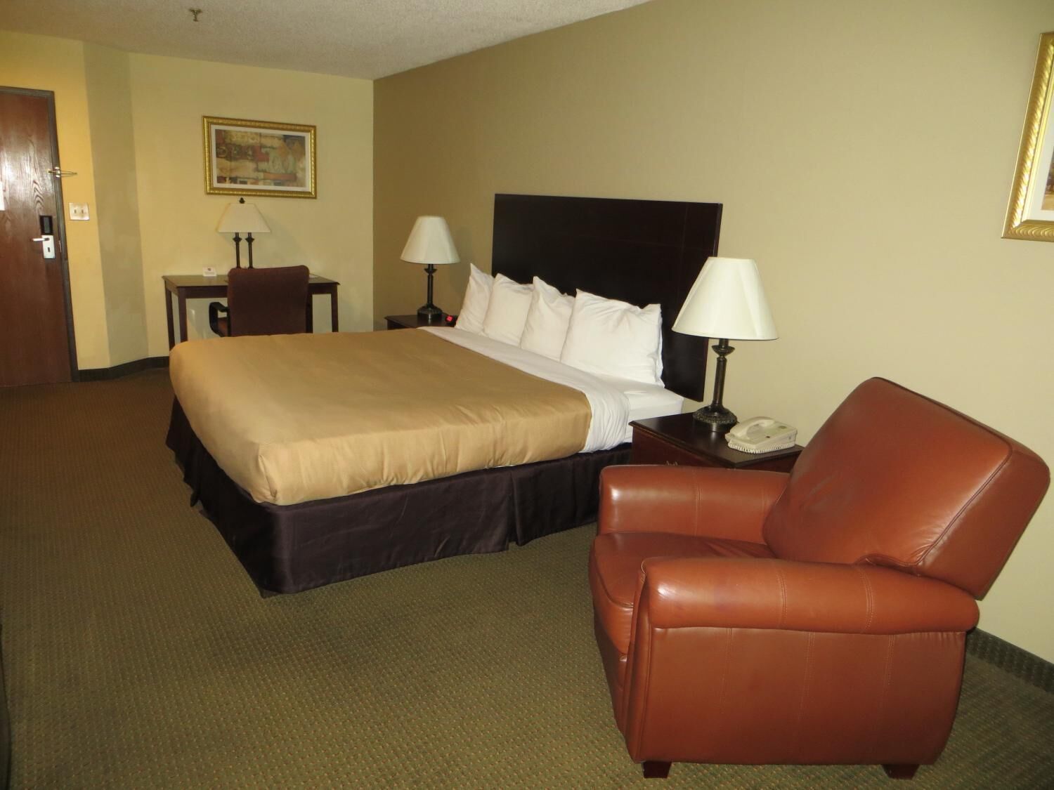 Quarto de hotel com uma cama grande, duas mesas de cabeceira com luminárias, uma poltrona de couro marrom, uma escrivaninha e uma cadeira.