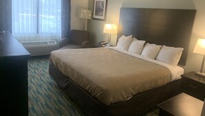 A hotel room with a large bed, a desk, a chair, a window with curtains, a wall-mounted lamp, and a framed picture.