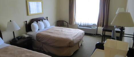 A hotel room with two beds, a desk, a chair, a lamp, a telephone, and an air conditioning unit.