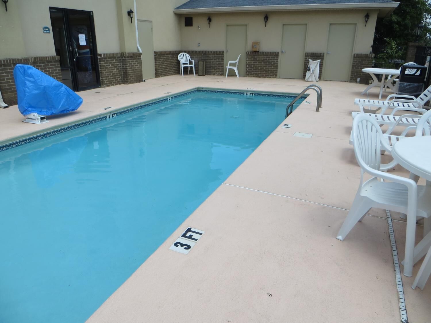 A rectangular pool with a 3 FT depth marker, surrounded by white plastic chairs and tables, and a building with a brick facade in the background.
