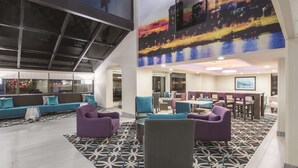 Een ruime lobby met een modern ontwerp, met een grote zithoek met paarse en grijze stoelen, een kamerbreed tapijt en een stadslandschap muurschildering.