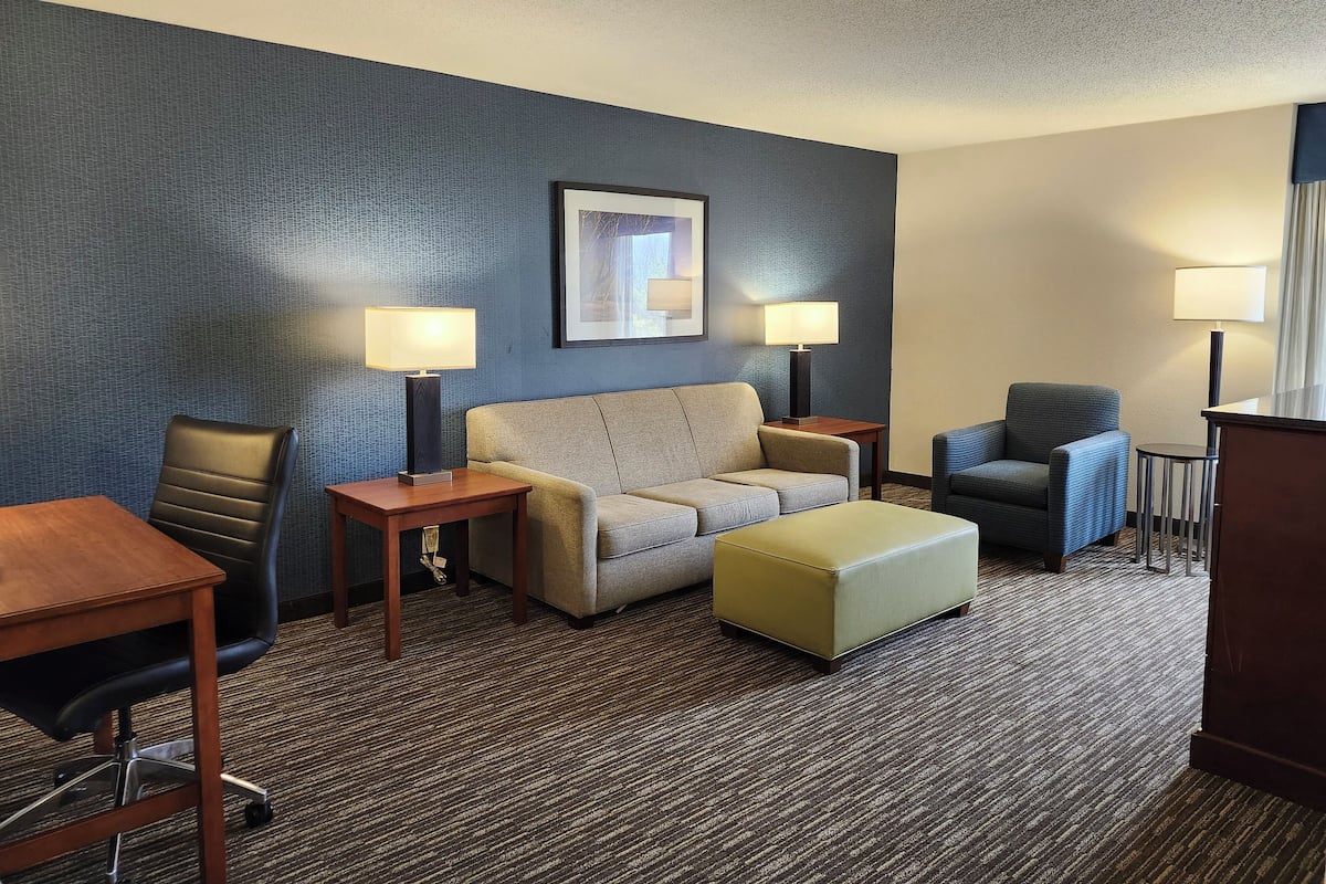 A hotel room with a sofa, armchair, ottoman, desk, and lamps.