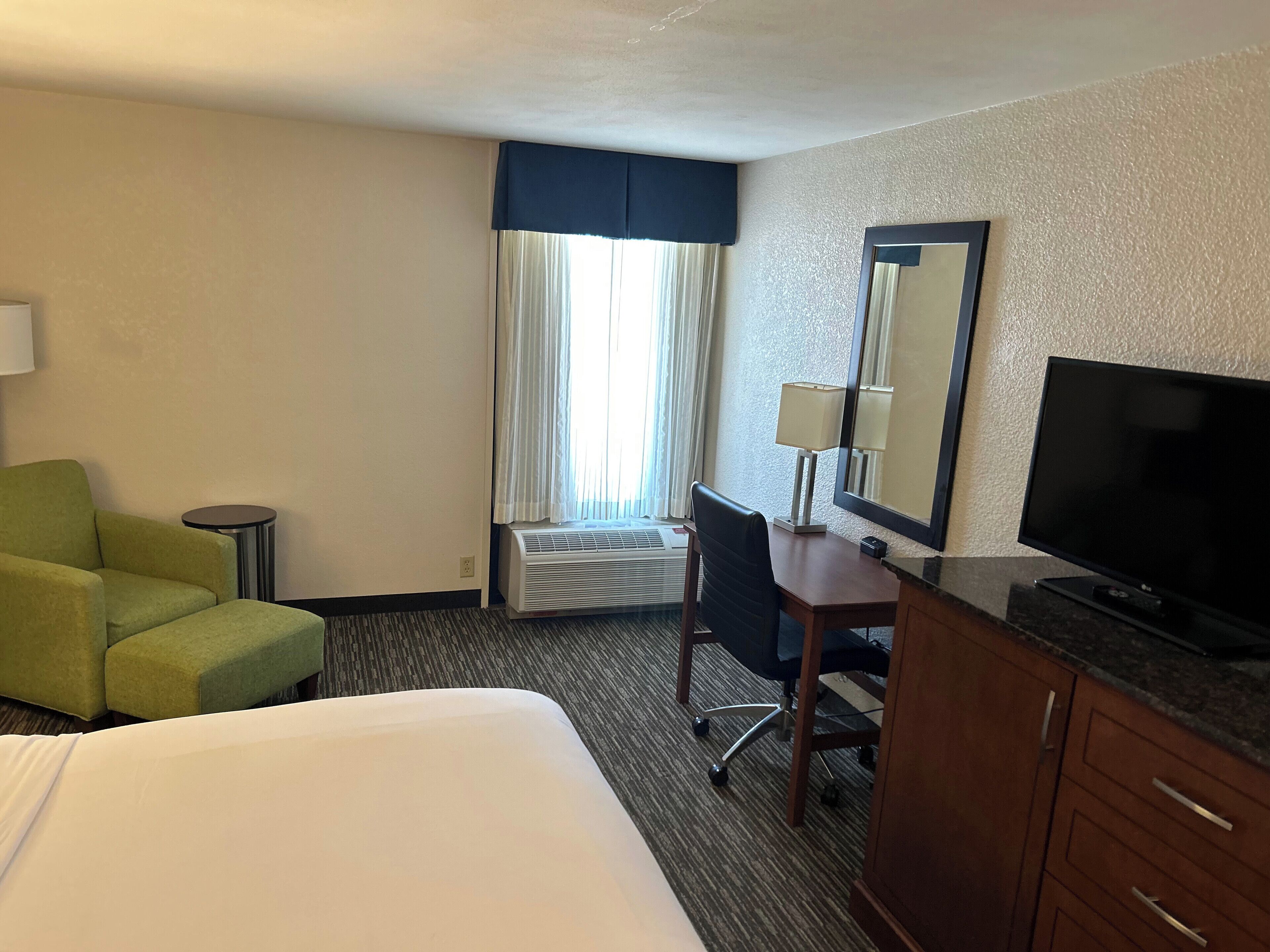 A hotel room with a bed, a desk, a chair, a television, a lamp, and a window with curtains.