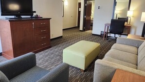 A hotel room with a flat-screen TV, a wooden cabinet, a light-colored sofa, a matching ottoman, and a small table. - Heritage Inn & Suites St. Louis/Fenton, Trademark by Wyndham (Fenton)