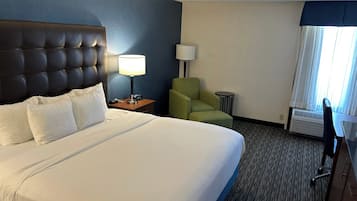 A hotel room with a large bed, a headboard, a bedside table with a lamp, a green armchair, a small round table, and a desk with a chair.