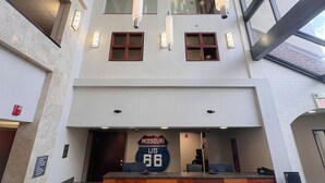 A modern hotel lobby with a high ceiling, multiple hanging pendant lights, a reception desk with a Missouri US 66 sign, and large windows allowing natural light. - Heritage Inn & Suites St. Louis/Fenton, Trademark by Wyndham (Fenton)