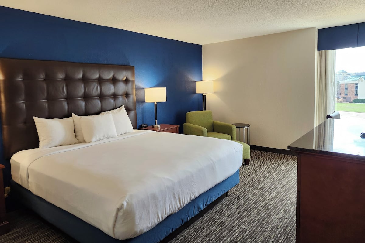 A hotel room with a large bed, two bedside tables, a green armchair, and a view of the outside.