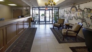 Un hall de hotel con mostrador de recepción, dos sillones, una alfombra estampada y un muro de mosaicos.