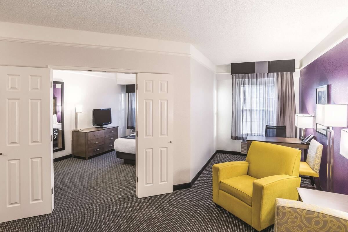 Une chambre d’hôtel avec un lit, un bureau et une chaise, un fauteuil jaune, une télévision et une salle de bain visible à travers une porte ouverte.