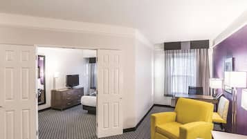 A hotel room with a bed, a desk with a chair, a yellow armchair, a TV, and a bathroom visible through an open door.
