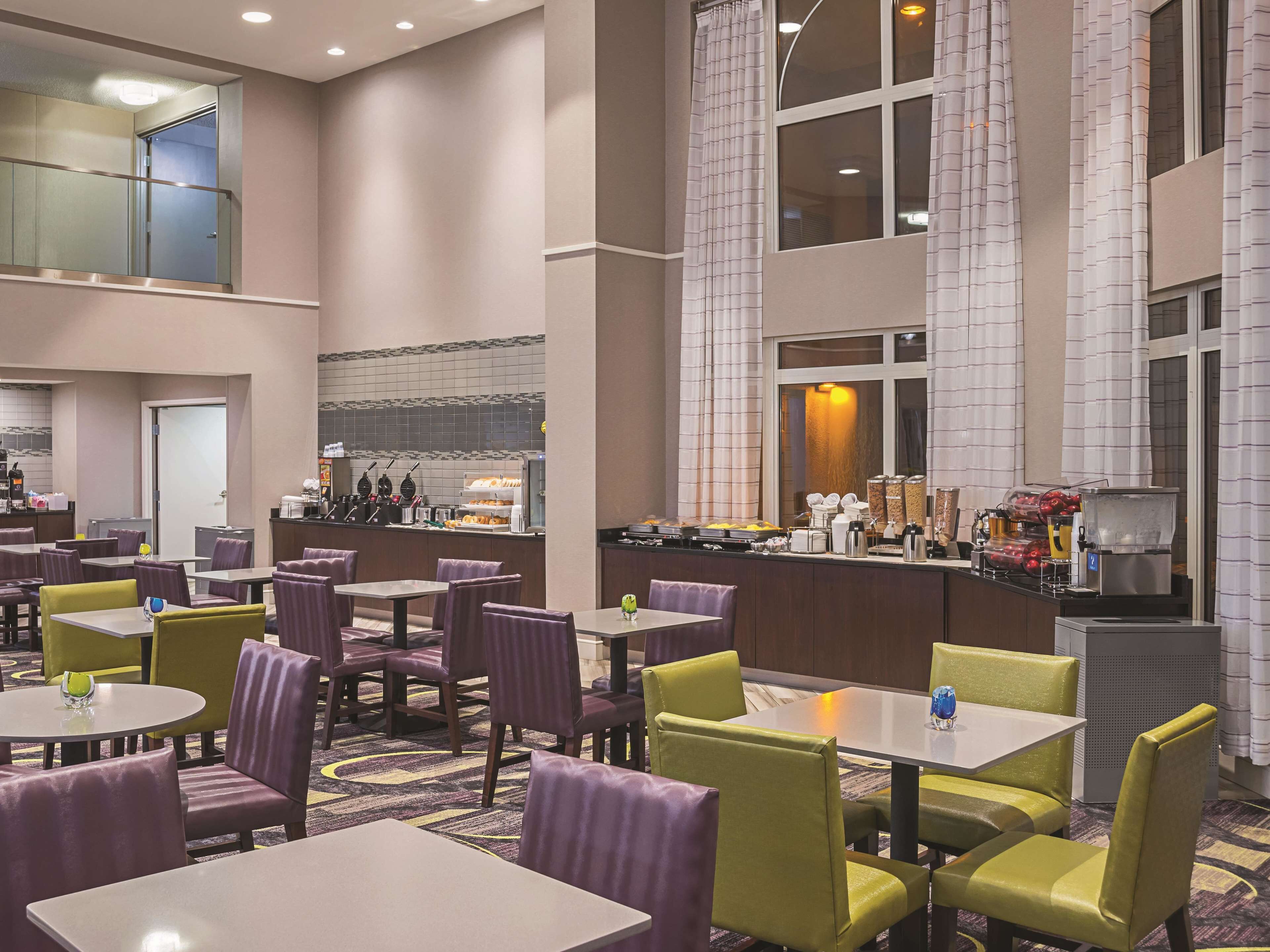 A hotel lobby with multiple dining tables and chairs, a kitchen area, and a large window with curtains.