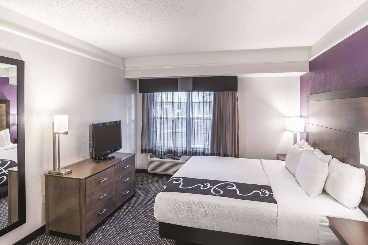 Une chambre d’hôtel équipée d’un lit, d’une télévision, d’une commode, d’un miroir et d’une fenêtre avec des rideaux.