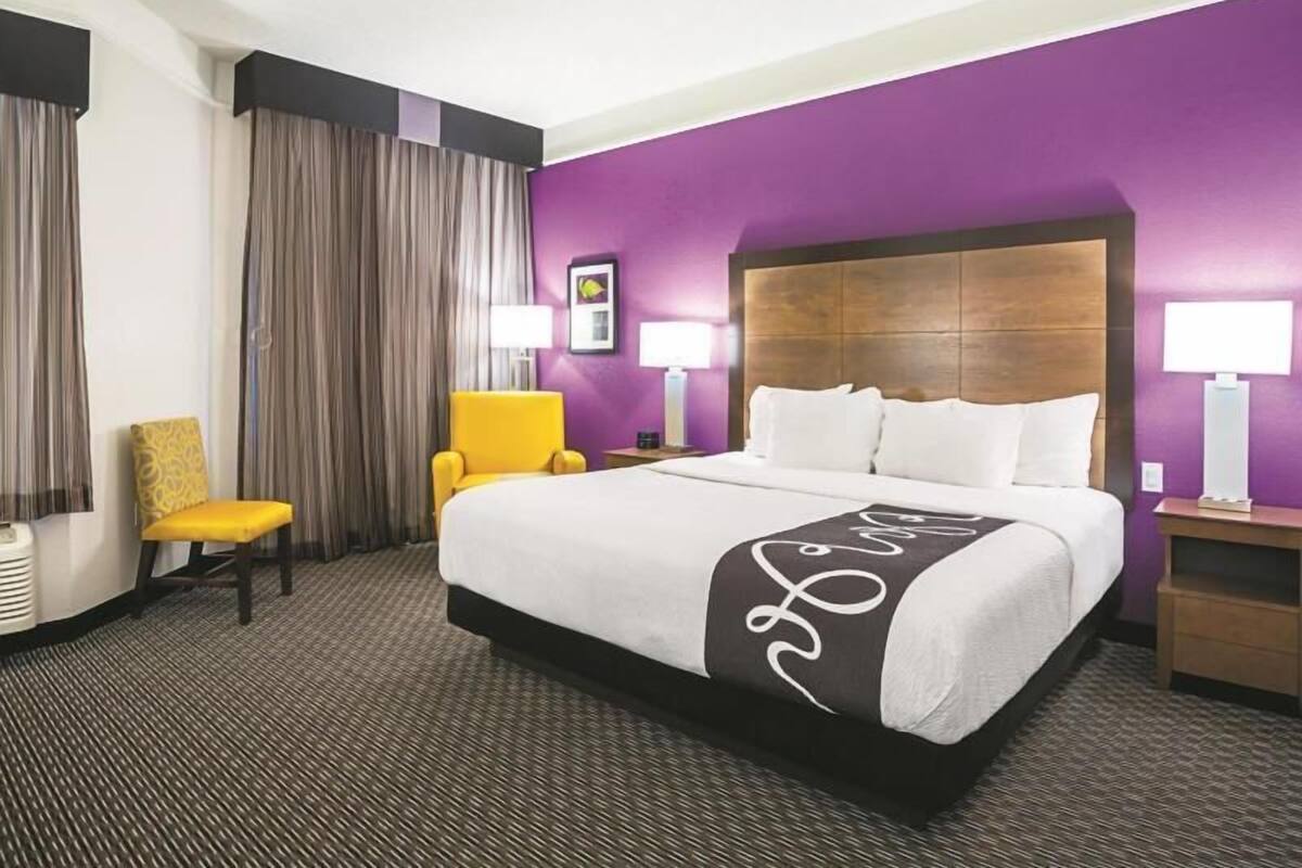 Une chambre d’hôtel avec un grand lit, un fauteuil jaune, une table de chevet avec une lampe et une vue sur l’extérieur.
