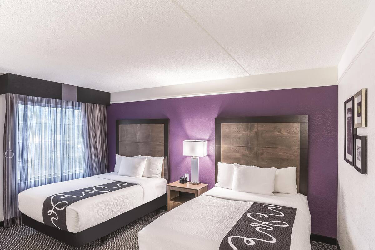 Une chambre d’hôtel avec deux lits, une table de chevet avec une lampe et des murs violets.