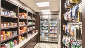A grocery store aisle with shelves stocked with snacks and beverages, a refrigerated display case, and a wall with hanging packaged goods.