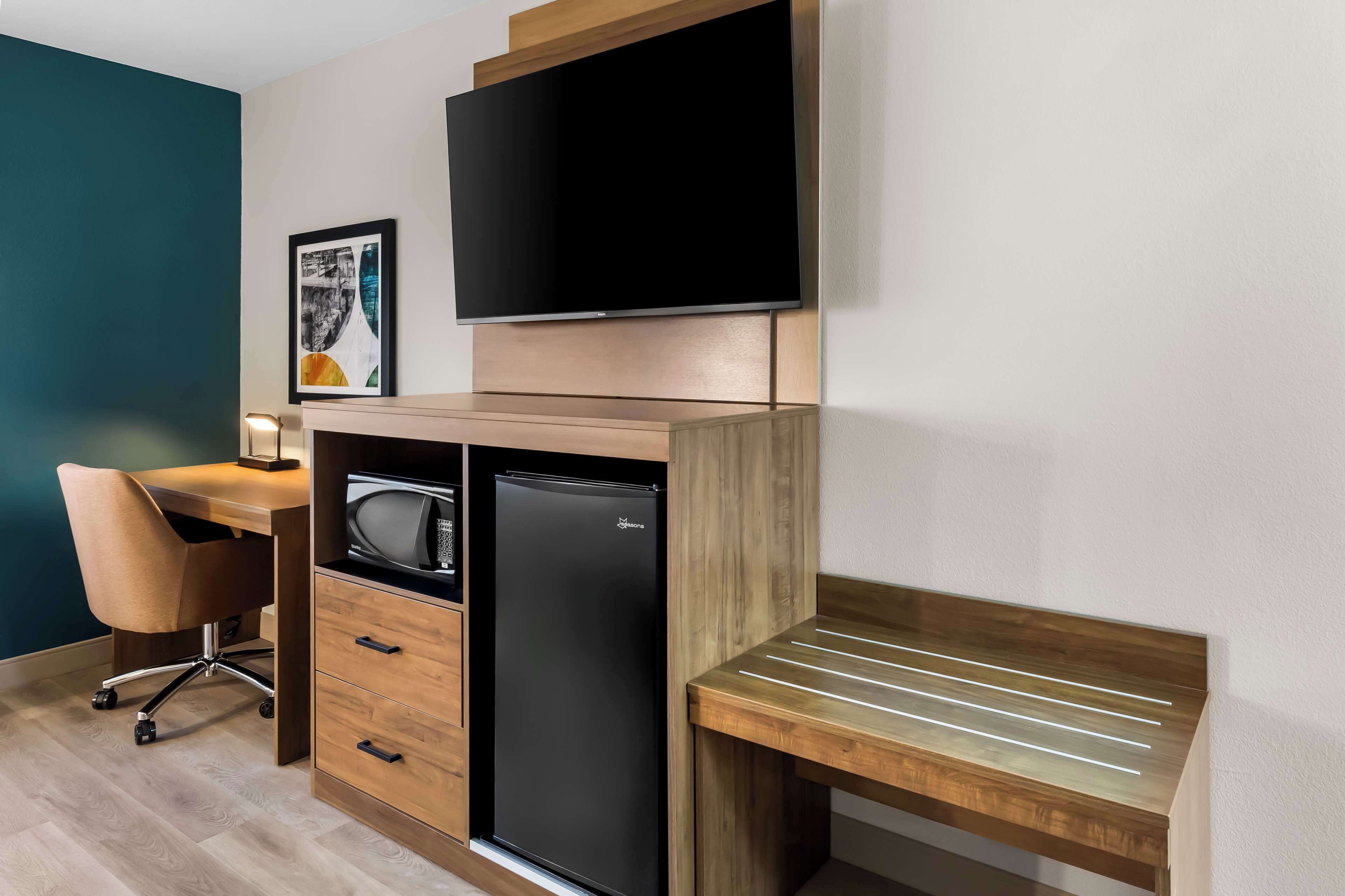 A compact hotel room with a wooden desk, a flat-screen TV, a refrigerator, a chair, and a bedside table.
