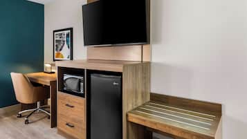 A compact hotel room with a wooden desk, a flat-screen TV, a refrigerator, a chair, and a bedside table.