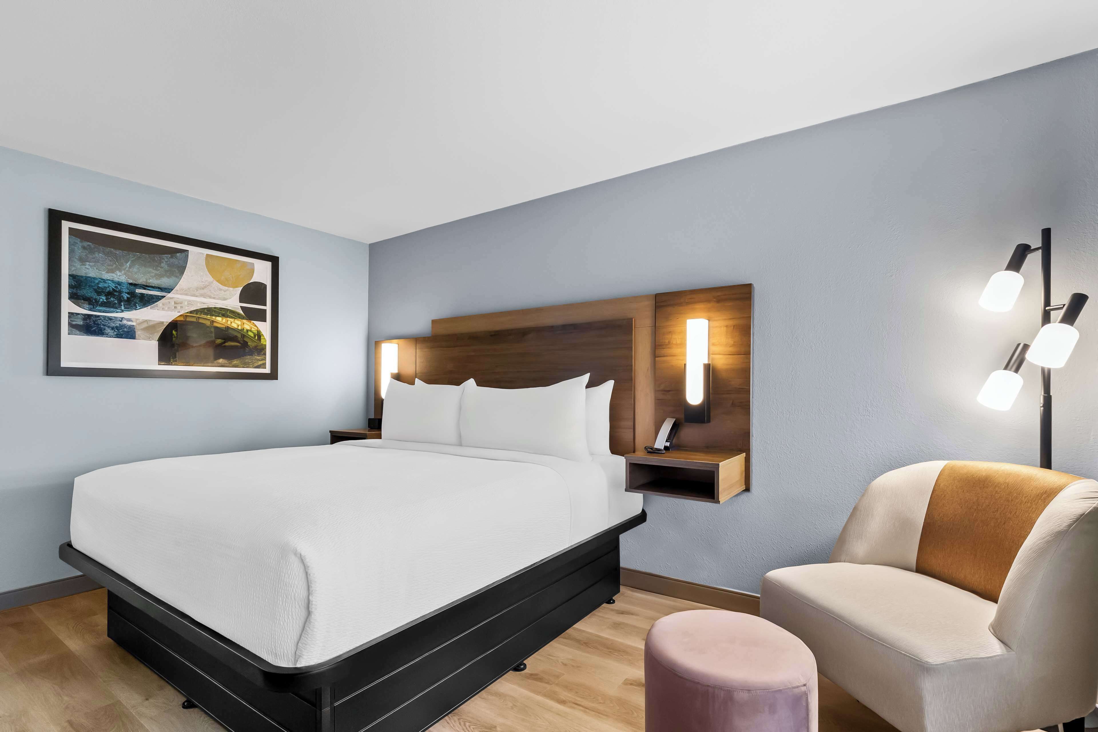 Une chambre d’hôtel moderne avec un grand lit, une tête de lit en bois, une table de chevet avec une lampe et une œuvre d’art accrochée au mur.