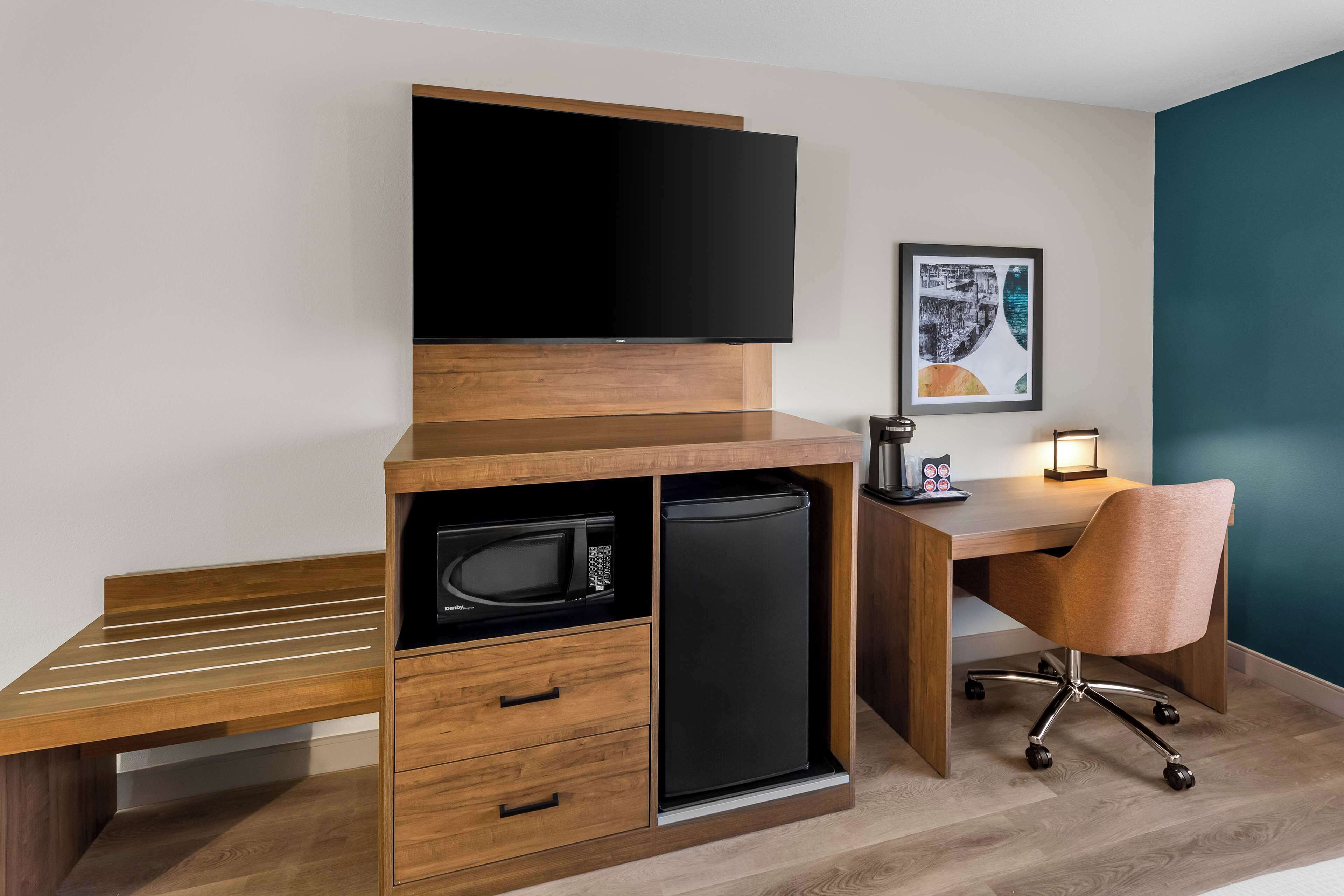 Una habitación de hotel compacta con un escritorio de madera, un televisor de pantalla plana, un microondas, un refrigerador, una silla acolchada y un cuadro en la pared.