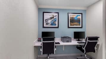 Una oficina moderna con dos escritorios, cada uno con un monitor de computadora y teclado. Hay dos cuadros enmarcados en la pared, una impresora y una lámpara de escritorio.
