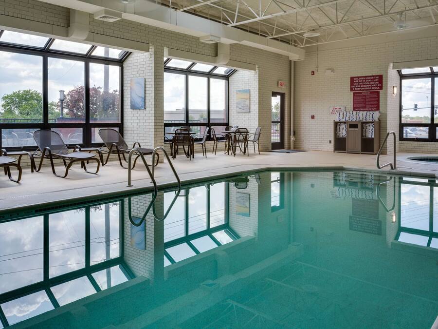 An indoor swimming pool with clear glass walls, a tiled floor, and a seating area with chairs and tables.