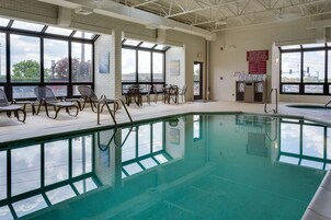 Una piscina interior con paredes de vidrio transparente, suelo de baldosas y una zona de estar con sillas y mesas.