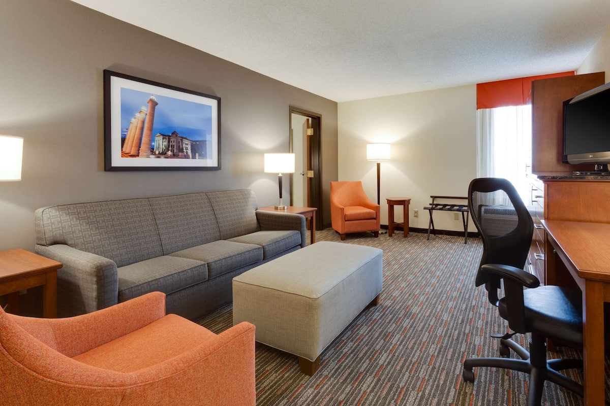 Habitación de hotel con un sofá gris, un sillón naranja, un pequeño otoman, un escritorio con computadora, televisor y un cuadro en la pared.