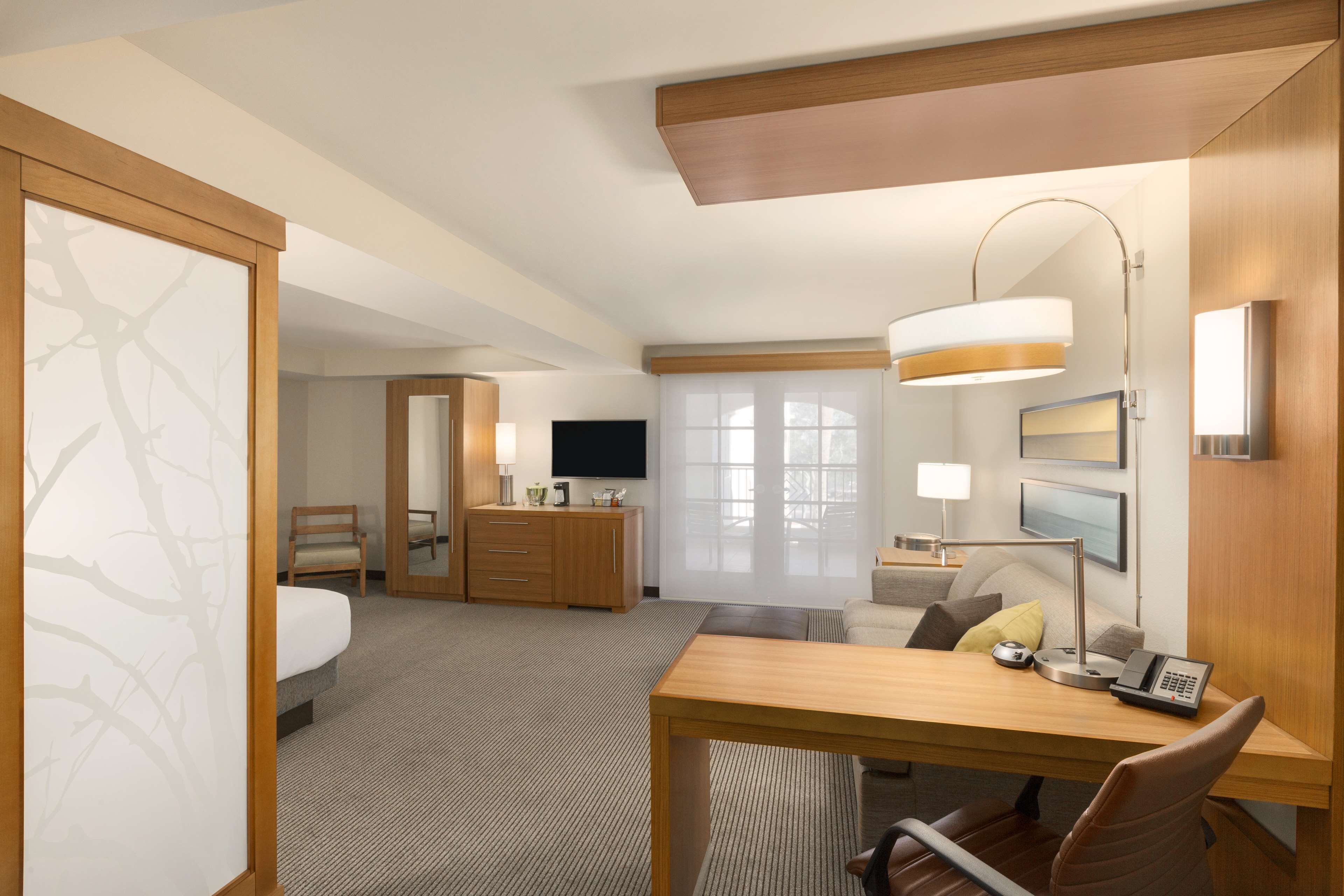 A hotel room with a bed, desk, chair, lamp, TV, and a view of the outdoors.