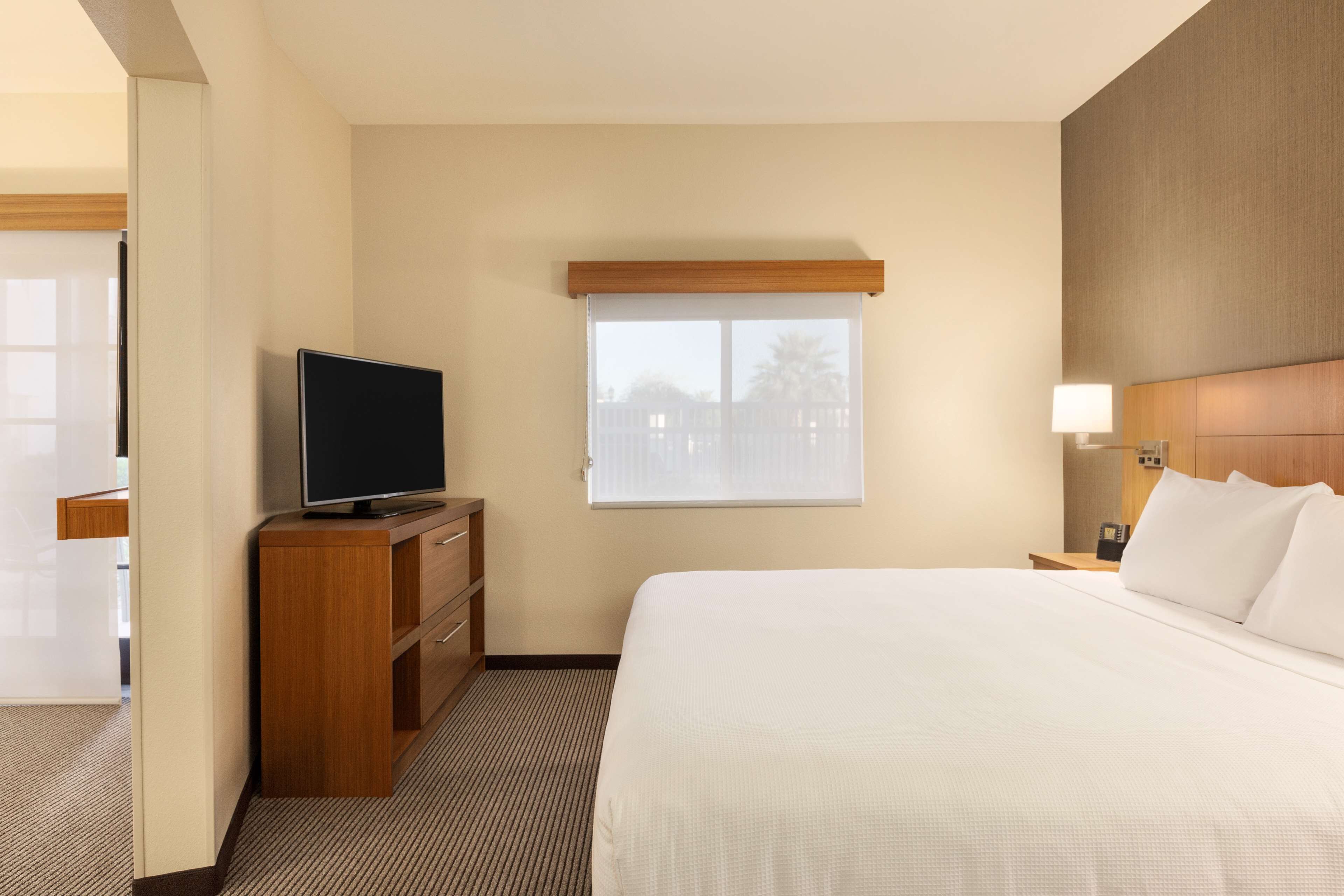 Habitación de hotel con una cama, un televisor sobre un aparador de madera, una ventana con persianas, una mesita de noche con lámpara y un cabecero de madera.