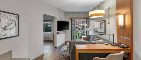 A modern hotel room with a wooden desk, a comfortable sofa, a flat-screen TV, and a large window offering a view of the outside.