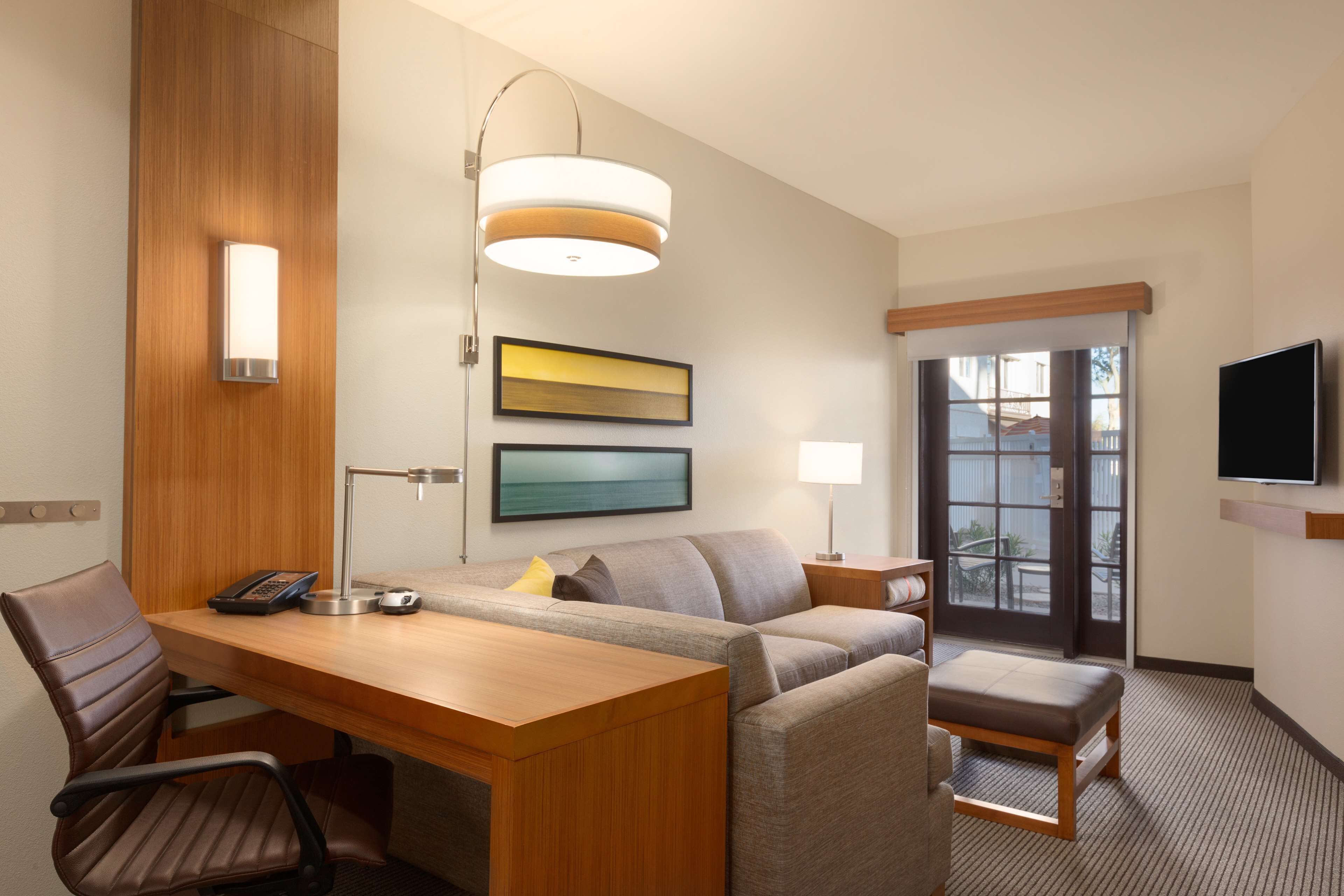 A hotel room with a wooden desk, a sofa, a small table, a chair, a lamp, a TV, and a window with a view.