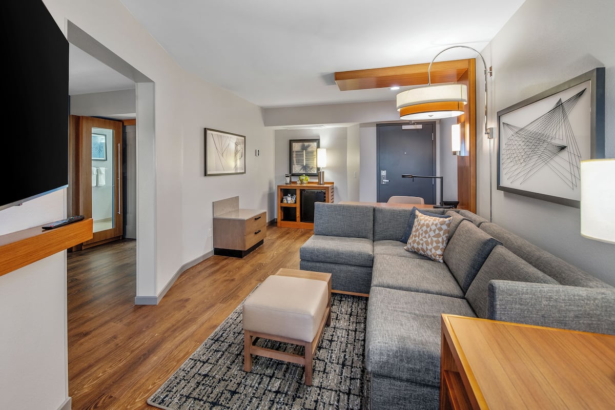 A modern living room with a sectional sofa, a wooden coffee table, and a rug. There is a small side table and a lamp. The room has wooden flooring and a door leading to another area.