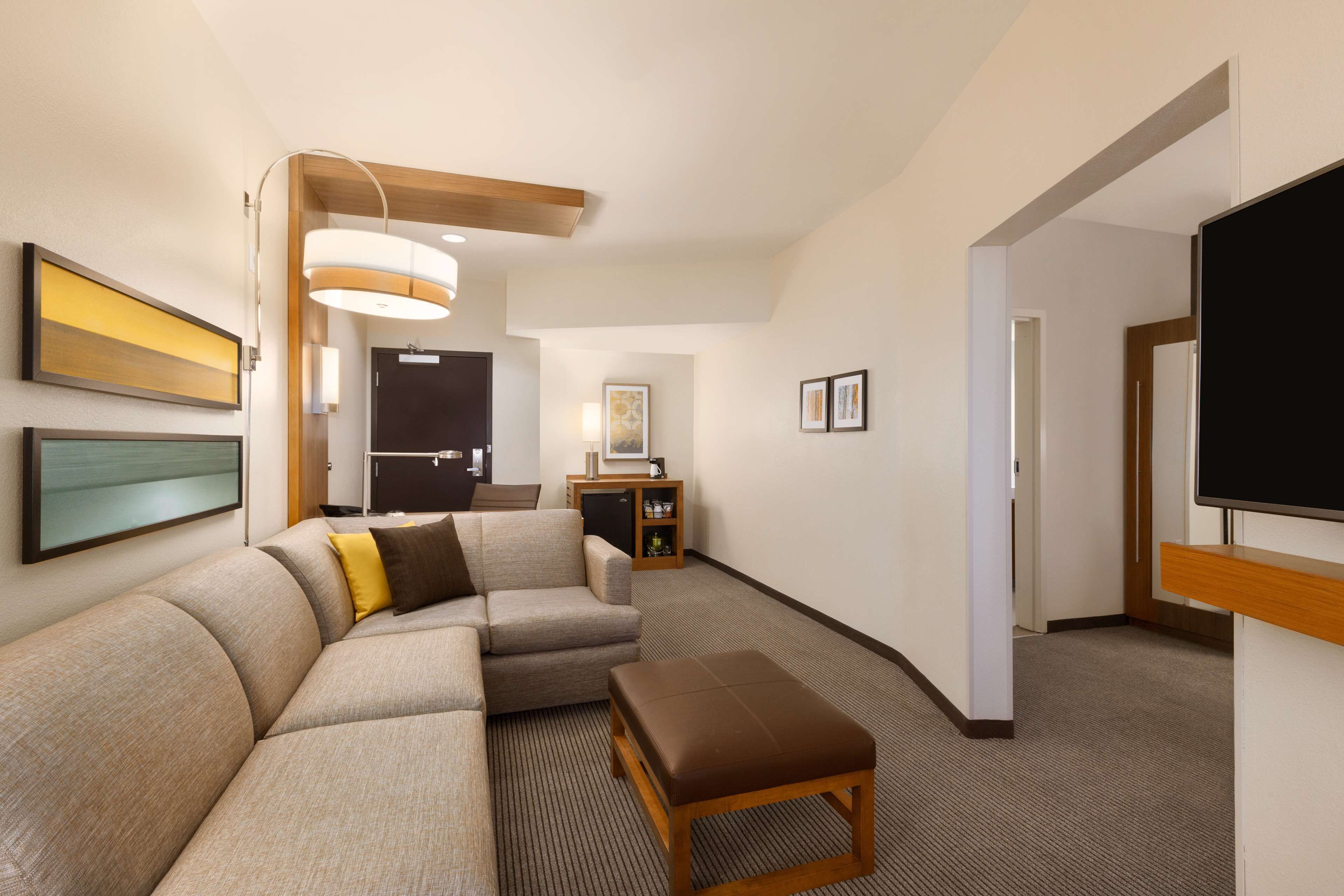 A modern hotel room with a sectional sofa, a small wooden table, a flat-screen TV, and a kitchenette area.