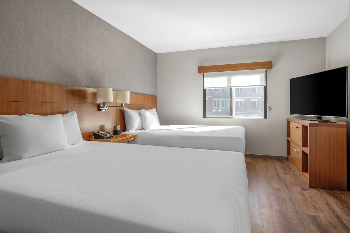 A hotel room with two beds, a TV, a desk, and a window with a view of buildings.