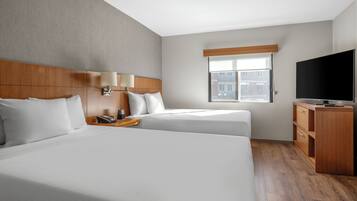 A hotel room with two beds, a TV, a desk, and a window with a view of buildings.