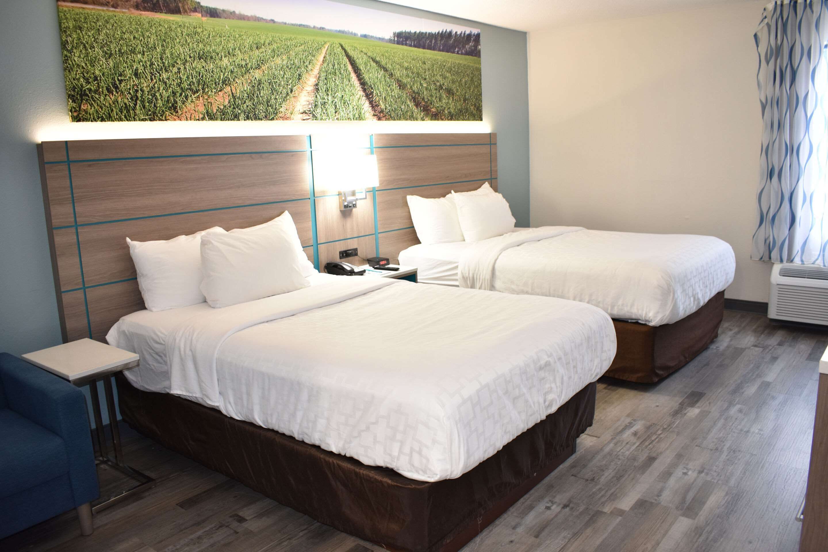 Habitación de hotel con dos camas, una mesita de noche, una pequeña mesa y un ventanal con vistas a un campo.