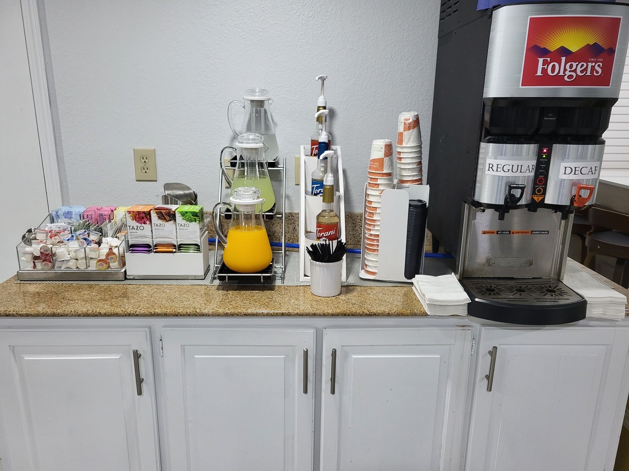 Un comptoir de cuisine avec une machine à café, une carafe de jus d’orange et divers condiments.