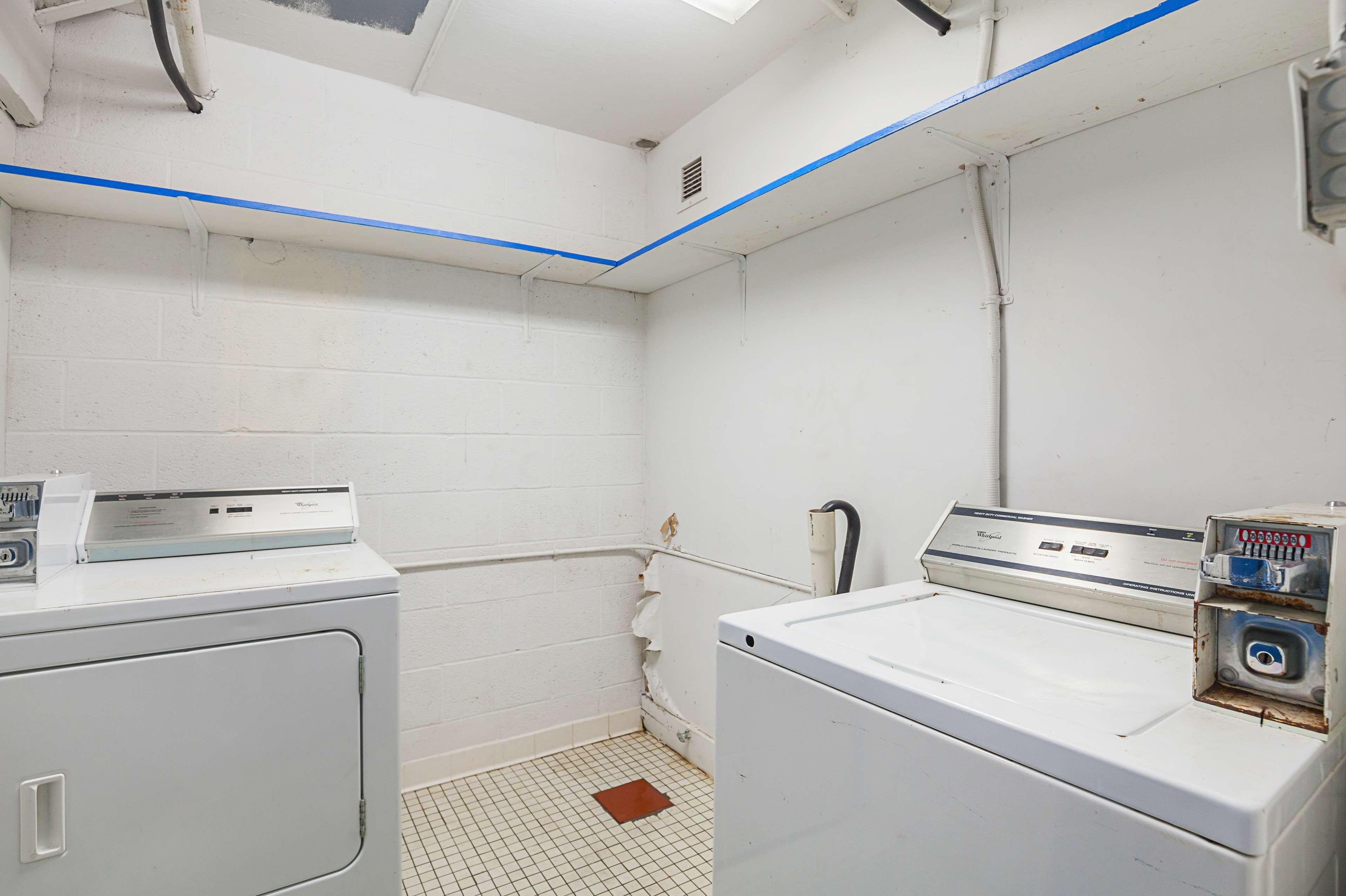 A laundry room with two white washers and dryers, white tiled walls, and a grid-patterned floor.