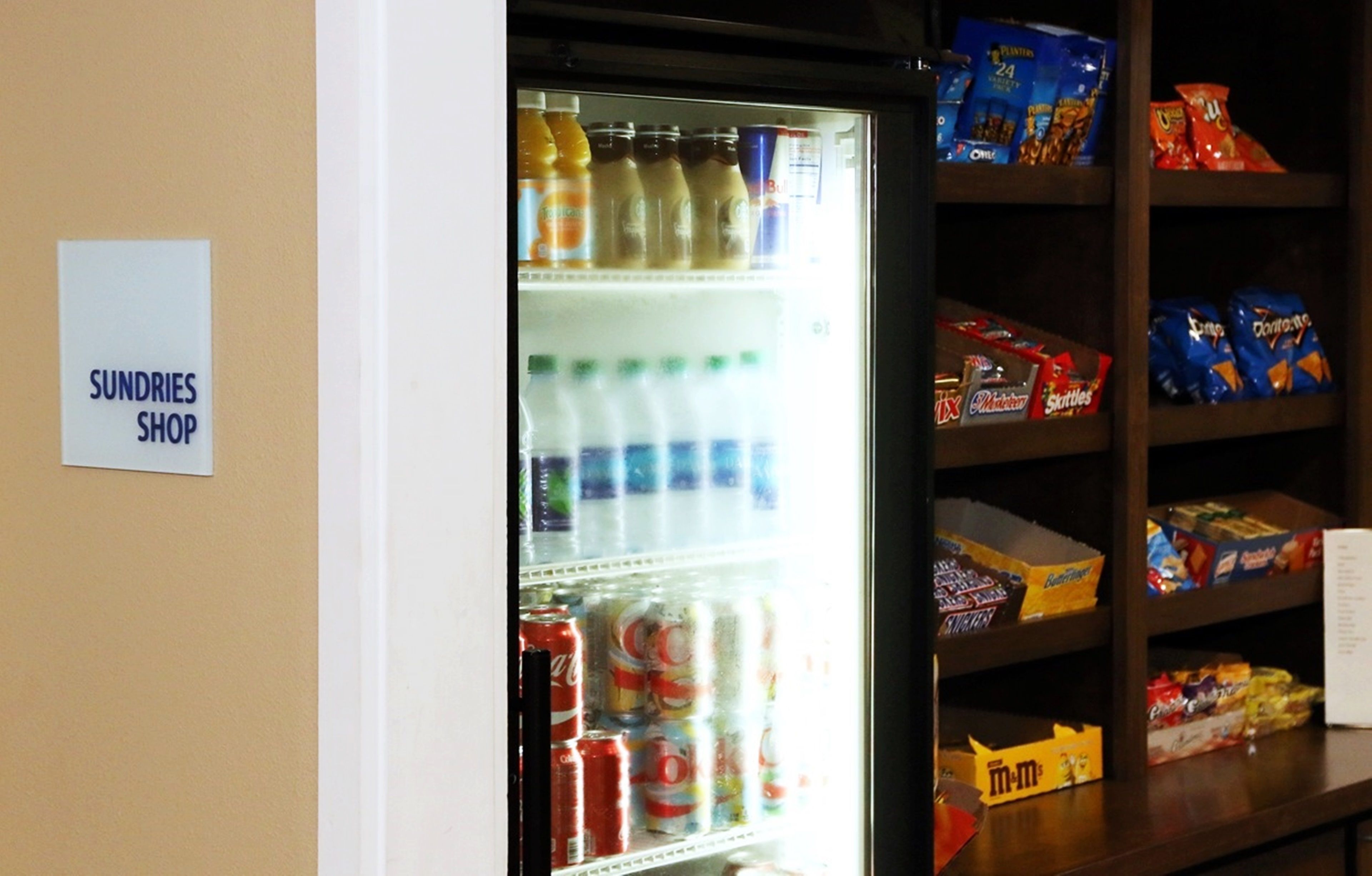 A refrigerator stocked with beverages and snacks, a sign reading "SUNDRIES SHOP", and shelves with packaged goods.