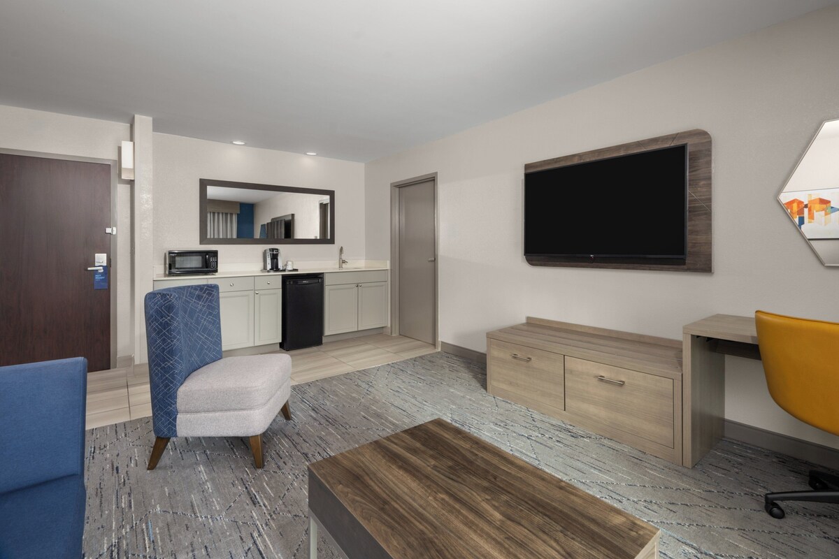 A modern hotel room with a kitchenette, a large flat-screen TV, a wooden desk, a yellow office chair, a blue armchair, and a small sofa.