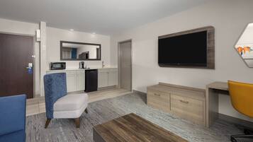 A modern hotel room with a kitchenette, a large flat-screen TV, a wooden desk, a yellow office chair, a blue armchair, and a small sofa.